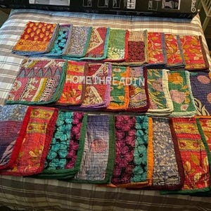 Può includere: Una collezione di pezzi di tessuto colorati e fantasia, probabilmente trapunte o coperte, disposti su un letto. I tessuti presentano una varietà di disegni, tra cui motivi floreali e geometrici, nelle tonalità del rosso, blu, verde e arancione. Il testo "HOMETHREADIN" è visibile.