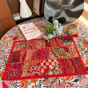 Könnte beinhalten: Ein rechteckiger Patchwork-Textil in Rot-, Gold- und Cremetönen mit rotem Rand und Quasten. Der Textil weist verschiedene Muster und Texturen auf, darunter Blumen- und geometrische Designs. Ein Buch mit Text ist sichtbar.