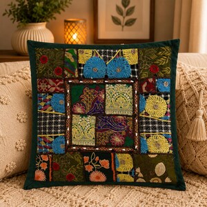 May include: A square throw pillow featuring a patchwork design of various embroidered fabrics in shades of dark green, blue, yellow, and red. The center panel displays intricate floral and geometric patterns, framed by a dark green border.
