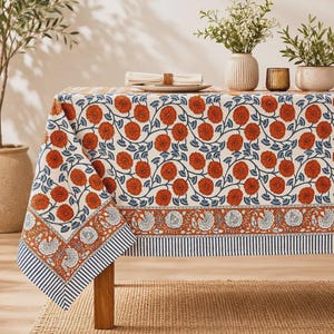 Puede incluir: Un mantel rectangular con un patrón repetitivo de flores naranjas con tallos y hojas azules sobre un fondo crema. El borde presenta una banda horizontal de motivos florales naranjas y blancos con un remate de rayas azul marino y blancas.