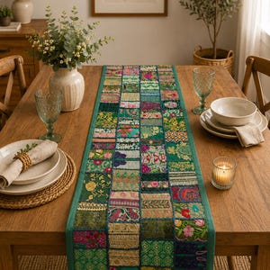 Puede incluir: Un camino de mesa rectangular con un diseño de patchwork de varios cuadrados de tela en tonos verdes, rosas y dorados. El camino está compuesto por múltiples segmentos textiles bordados y estampados unidos con un borde verde sólido.