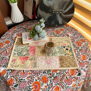 Op de afbeelding: Een ronde tafel bedekt met een tafelkleed met bloemenpatroon. Een patchwork placemat met kwastjes ligt erop, versierd met bloemen- en geborduurde details. Een kleine mozaïeken kandelaar en een boek staan ook op de tafel.