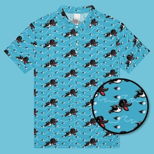 Puede incluir: Polo de color azul claro con un estampado repetido de patos de dibujos animados, pelotas de golf y líneas onduladas. Los patos son negros y blancos con detalles en rojo. La camisa tiene cuello y mangas cortas. La manga tiene el texto "Club Carts".