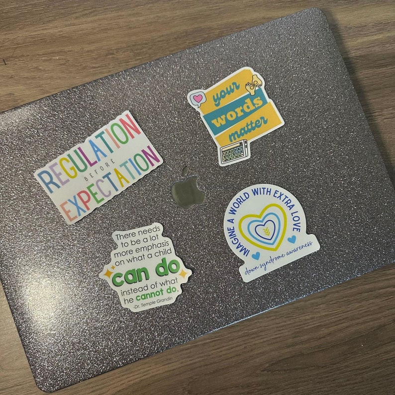 May include: A laptop computer with several colorful stickers on it. The stickers have inspirational messages such as "Regulation Before Expectation", "Your Words Matter", "Imagine a World with Extra Love", and "There needs to be a lot more emphasis on what a child can do instead of what he cannot do. Dr. Temple Grandin".