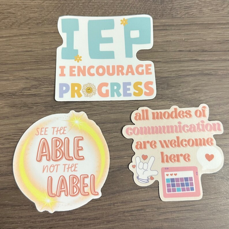 May include: Three colorful stickers with positive messages. The first sticker is blue and pink and says "IEP I encourage progress". The second sticker is yellow and says "See the able not the label". The third sticker is pink and says "All modes of communication are welcome here".