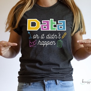 以下が含まれることがあります： カラフルなブロック文字で「Data or it didn't happen」と書かれたダークグレーのTシャツ。テキストは、鉛筆、チェックリスト、グラフなど、カラフルなアイコンで囲まれています。
