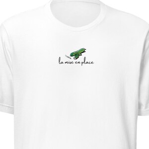 May include: White t-shirt with a small green and white graphic of a cutting board and knife, with the text "la mise en place" in black script. The shirt is made of a soft material.