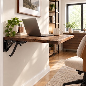 May include: A wall-mounted, fold-down desk featuring a dark brown wood grain surface with a live edge. The desk is supported by black metal folding brackets attached to the wall. A silver laptop, a potted green plant, and a mug sit on the desk.