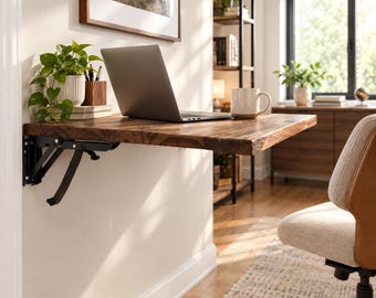 May include: A wall-mounted, fold-down desk featuring a dark brown wood grain surface with a live edge. The desk is supported by black metal folding brackets attached to the wall. A silver laptop, a potted green plant, and a mug sit on the desk.