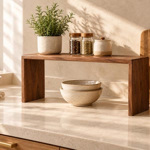 May include: A rectangular dark wood countertop shelf with a smooth finish and two vertical side supports. The shelf holds a speckled ceramic pot with a green plant, two glass spice jars with wooden lids, and a small ceramic container with a lid.