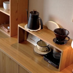 Könnte beinhalten: Ein hölzernes Regalorganizer mit einem schwarzen Schwanenhalskessel auf einem weißen Untersetzer, einem schwarzen Kaffeefilterhalter, einer cremefarbenen Keramiktasse mit Untertasse, einem weißen Papierfilter und einer schwarzen Digitalwaage.