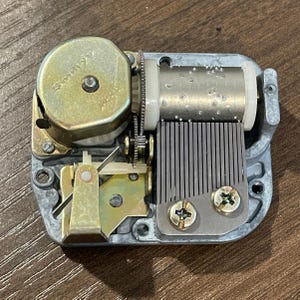 May include: A silver-colored music box mechanism with a brass winding key and a metal comb. The mechanism has a white plastic cylinder with a series of holes and a brass screw. The text "Sonora" is visible on the winding key.