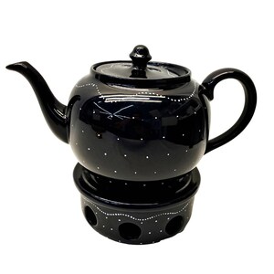 May include: A glossy black ceramic teapot with a matching warmer base, both decorated with hand-painted white dots and wavy lines. The teapot features a curved spout, a rounded handle, and a lid with a small knob, while the base has circular cutouts.