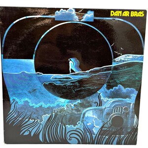 May include: Vintage vinyl record album cover with a blue and black illustration. The artwork depicts a figure standing on a rocky shore, with a large arch and clouds in the background. The album title "DAN AR BRAS" is in yellow text.
