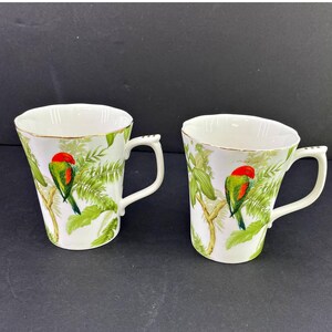 May include: Two white ceramic mugs featuring a vibrant tropical pattern of green palm leaves and branches with a small parrot perched on each. The parrots have bright red heads and green bodies. Each mug has a gold-trimmed rim and a white handle.