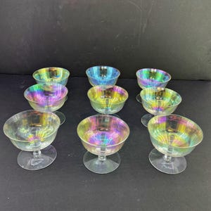 May include: A set of eight glass dessert bowls featuring a translucent, iridescent finish that reflects rainbow colors. Each bowl has a wide, shallow basin supported by a short, clear glass stem and a circular base, arranged in a grid pattern.