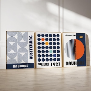 Op de afbeelding: Drie ingelijste Bauhaus kunstprints. De prints tonen geometrische ontwerpen in marineblauw, oranje en wit. De tekst "Bauhaus" en "1923" is zichtbaar. De frames zijn lichtgoud van kleur.