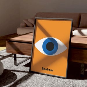 Puede incluir: Un p&oacute;ster enmarcado con un fondo naranja brillante y un gran ojo azul con una pupila negra. El texto "Bauhaus" est&aacute; impreso en negro en la parte inferior del p&oacute;ster.