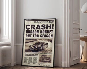 May include: A framed vintage-style newspaper print with the headline "CRASH! HUDSON HORNET OUT FOR SEASON." The print features a sepia-toned image of a classic car and additional text. The frame is black, and the print is displayed against a neutral wall.