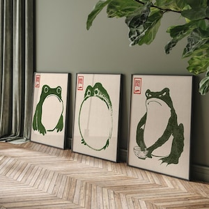 May include: Three framed prints of green frogs with a Japanese character in red on each print. The frogs are in different poses, one is sitting, one is holding a bowl, and one is standing with its mouth open.