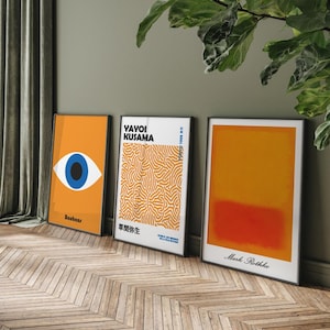 Puede incluir: Tres impresiones de arte enmarcadas con un dise&ntilde;o minimalista. La impresi&oacute;n de la izquierda presenta un ojo azul sobre un fondo amarillo con el texto "Bauhaus". La impresi&oacute;n del medio presenta un patr&oacute;n abstracto blanco y naranja con el texto "Yayoi Kusama" y texto japon&eacute;s. La impresi&oacute;n de la derecha presenta un degradado amarillo y naranja con el texto "Mark Rothko".