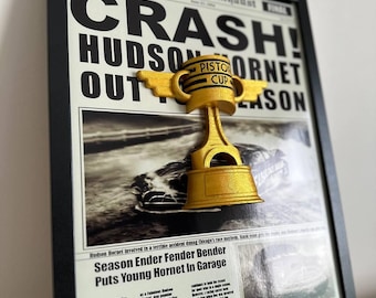 May include: A framed newspaper clipping with a gold trophy in the shape of a piston cup. The headline reads "CRASH! HUDSON HORNET OUT FOR SEASON". The article discusses a racing accident that ended the season for a young driver.