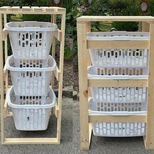 May include: A light-colored wooden storage rack featuring three stacked levels, each holding a white plastic laundry basket with a rectangular grid pattern. The frame is constructed from vertical and horizontal wooden slats, providing an open structure for the three removable baskets.