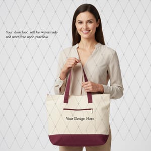 Peut inclure: Sac fourre-tout crème avec une base et des poignées bordeaux. Le sac a une poche avant zippée et le texte "Your Design Here" imprimé sur le devant. Le sac est tenu par une personne.