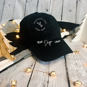 May include: A black baseball cap with the text "Your Design Here" on the front. The cap has a curved brim and a circular logo with text. The background includes decorative Christmas trees and string lights.