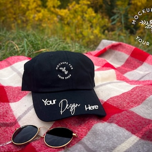 Może przedstawiać: Czarna czapka bejsbolówka z haftowanym białym logo z napisem "Mockups for your shop" i tekstem "Your Design Here" na daszku. Czapka leży na czerwono-białej kratkowanej narzucie.