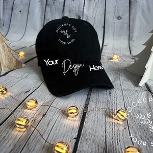 May include: Black baseball cap with white text that says "Mockups for Your Shop" and "Your Design Here". The cap is on a wooden surface with a string of lights and two small wooden Christmas trees.