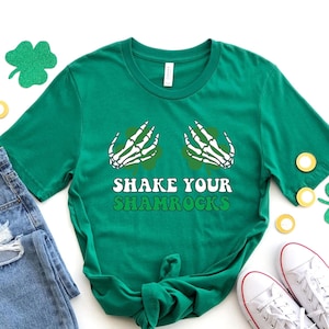 May include: A green t-shirt with a skeleton hand graphic holding a shamrock and the text "Shake Your Shamrocks". The shirt is laid out on a white surface with a pair of blue jeans, white trainers, and green shamrock decorations.