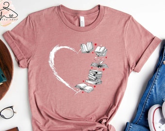 Pode incluir: Uma camiseta malva com um design de coração branco e uma pilha de livros com estrelas ao redor. A camiseta está colocada sobre uma superfície branca com um par de calças de ganga e ténis brancos.