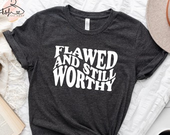 May include: A dark grey t-shirt with the white text "FLAWED AND STILL WORTHY" in a wavy font. The shirt is knotted at the hem. A neutral-toned hat, a cup of coffee, and a book are also in the image.