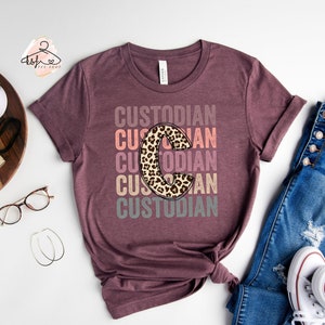 May include: A maroon t-shirt with the word "Custodian" repeated five times in different colours. The letter "C" in the word "Custodian" is a leopard print design.
