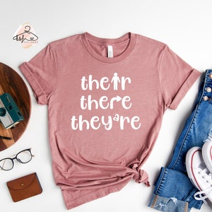 Puede incluir: Una camiseta color malva con las palabras "their there they're" impresas en blanco en la parte delantera.