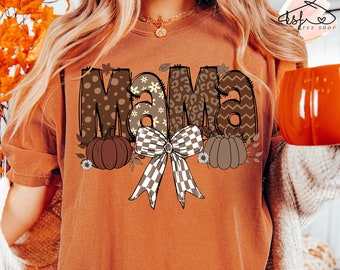 May include: A brown t-shirt with the word "Mama" in brown, orange, and white patterned letters. The letters are decorated with a brown bow, pumpkins, and fall-themed patterns.