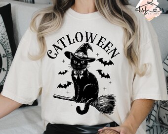 Könnte beinhalten: Ein weißes T-Shirt mit einer schwarzen Katze, die einen Hexenhut trägt und auf einem Besen reitet. Der Text "Catloween" steht über der Katze.