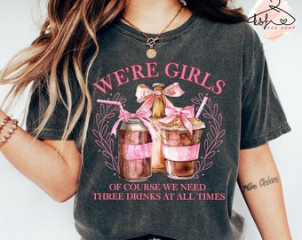 Puede incluir: Una camiseta gris con el texto "We're Girls" en letras rosas. La camiseta presenta una ilustración de acuarela de tres bebidas con lazos rosas y pajitas. El texto "Of course we need three drinks at all times" está impreso debajo de la ilustración.