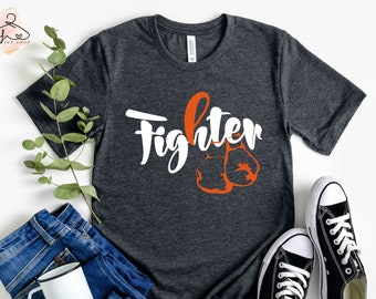 May include: Dark gray t-shirt with the word "Fighter" in white script and orange boxing gloves. The shirt is paired with blue jeans, a white mug, and black sneakers with white laces. A sprig of greenery adds a touch of freshness.