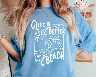 Puede incluir: Una camiseta azul claro con un gráfico blanco que dice "Life is Better at the Beach". El gráfico presenta una palmera, el sol y olas.