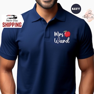 May include: Navy blue polo shirt with the text "Mrs. Ward" and a red apple graphic. The shirt has a collared neckline and short sleeves. The image also includes the word "Navy" in a circle and a logo for a tee shop.