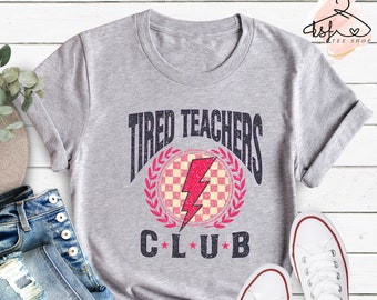 Könnte beinhalten: Graues T-Shirt mit einer abgenutzten Grafik, auf der "Tired Teachers Club" mit einem Blitz und einem karierten Muster in einem Kranz steht.