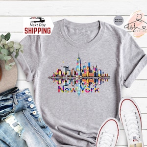 May include: A light gray t-shirt featuring a colorful graphic of the New York City skyline with the text "New York". The shirt is paired with blue jeans and white sneakers.