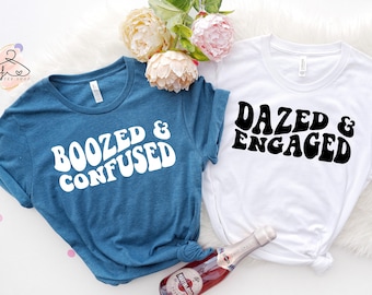 May include: Two t-shirts with a retro font design. One shirt is teal blue with the text "Boozed & Confused" in white. The other shirt is white with the text "Dazed & Engaged" in black.