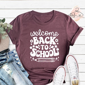 May include: A maroon t-shirt with the text "welcome back to school" in white. The text is in a retro font with stars and apples around it. A white pencil is drawn below the text. The shirt is laid out on a white wooden surface with a pair of white sneakers.