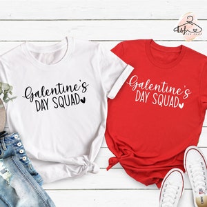 Puede incluir: Dos camisetas con el texto "Galentine's Day Squad" en letras blancas y negras sobre una camiseta blanca y roja. Las camisetas están dispuestas sobre una superficie de madera blanca con un par de zapatillas blancas.