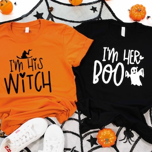 May include: Two orange and black Halloween themed t-shirts. The orange shirt says "I'm His Witch" with a witch's hat and heart. The black shirt says "I'm Her Boo" with a ghost.