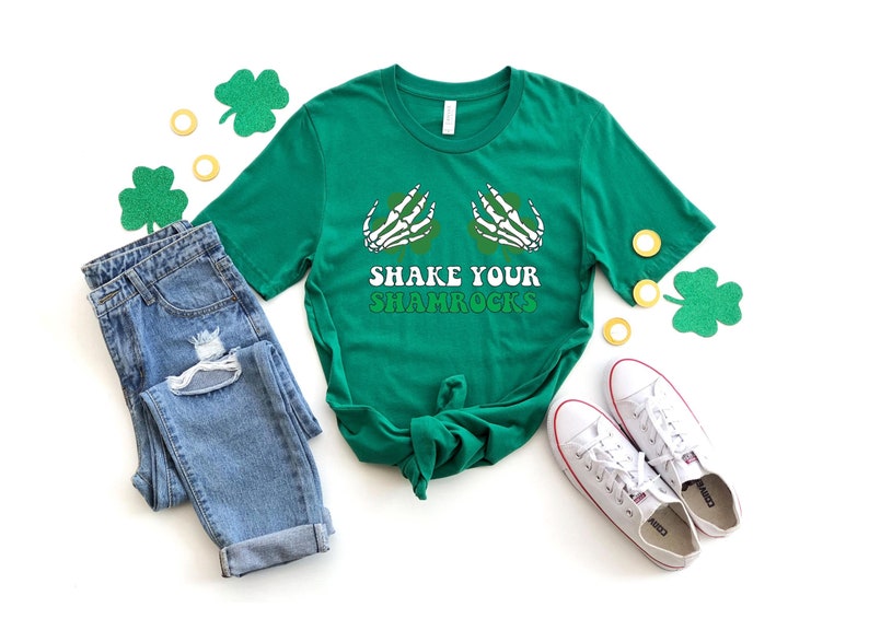 May include: A green t-shirt with a skeleton hand graphic holding a shamrock and the text "Shake Your Shamrocks". The shirt is laid out on a white surface with a pair of blue jeans, white trainers, and green shamrock decorations.
