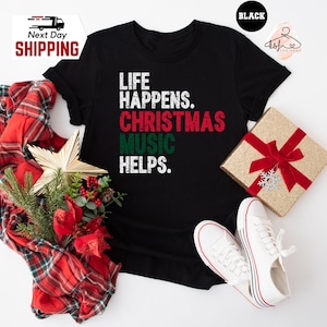 May include: Black t-shirt with the text "LIFE HAPPENS. CHRISTMAS MUSIC HELPS." in white, red, and green. The shirt is displayed with a wrapped gift, white sneakers, and a plaid scarf with Christmas decorations.
